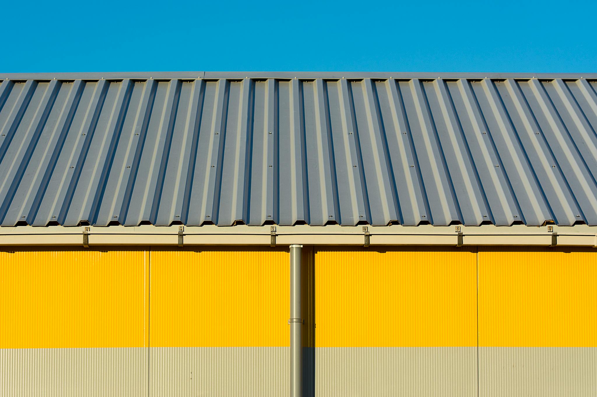 Photo of a yellow warehouse wall with a corrugated metal roof under a clear blue sky.